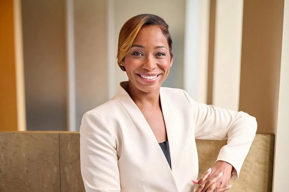 Portrait of Andrea Joy Campbell, a smiling Black woman with dark hair in an updo, wearing a cream blazer over a black top, seated against a warm-toned background.