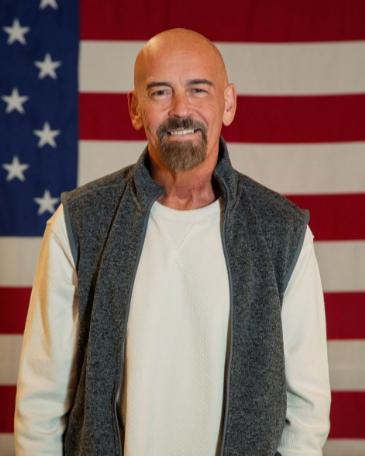 Portrait of John Deaton, a bald middle-aged white man with a goatee, wearing a gray fleece vest over a cream long-sleeve shirt, standing in front of an American flag.