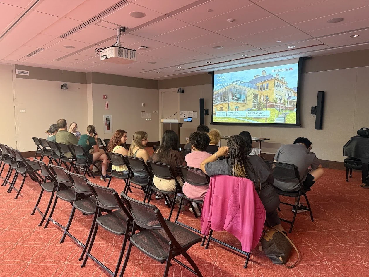 People sit in rows of folding chairs facing a projection screen in the library Community Room during the reception.