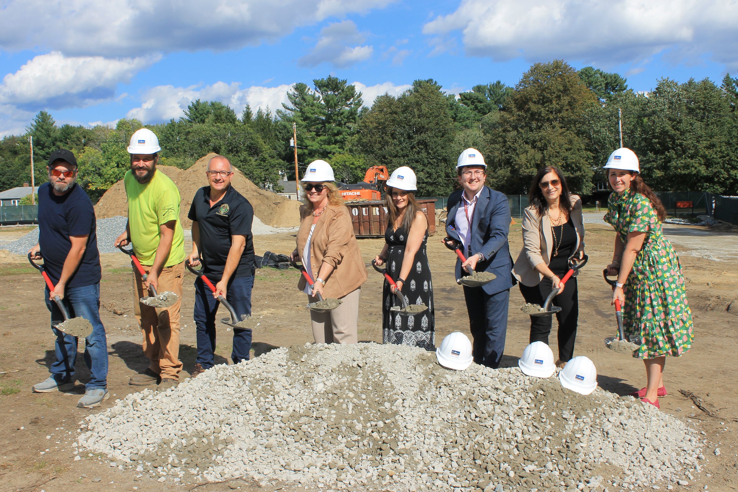 Eight people in white hard hats pose with ceremonial shovels over a pile of gravel at the Killam School groundbreaking site.