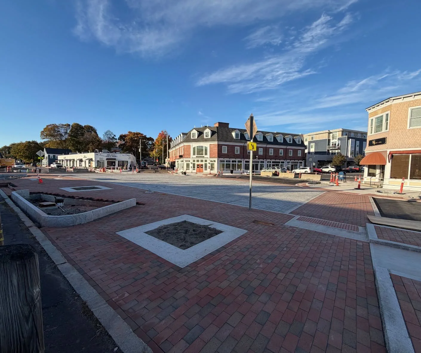 Brick sidewalks with granite-edged planting areas lead to a wide gray paved intersection with orange cones, a brick corner building, and a blue sky.