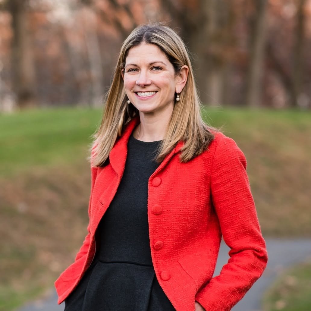 Portrait of Kate Lipper-Garabedian, a smiling woman with long blonde hair, wearing a red jacket over a black dress, standing outdoors in a park.