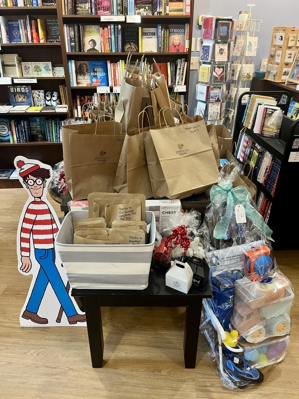 A cardboard Where's Waldo cutout stands beside a bookstore table piled with brown paper gift bags, cellophane-wrapped prize baskets, and handwritten thank-you notes, with book-lined shelves in the background.