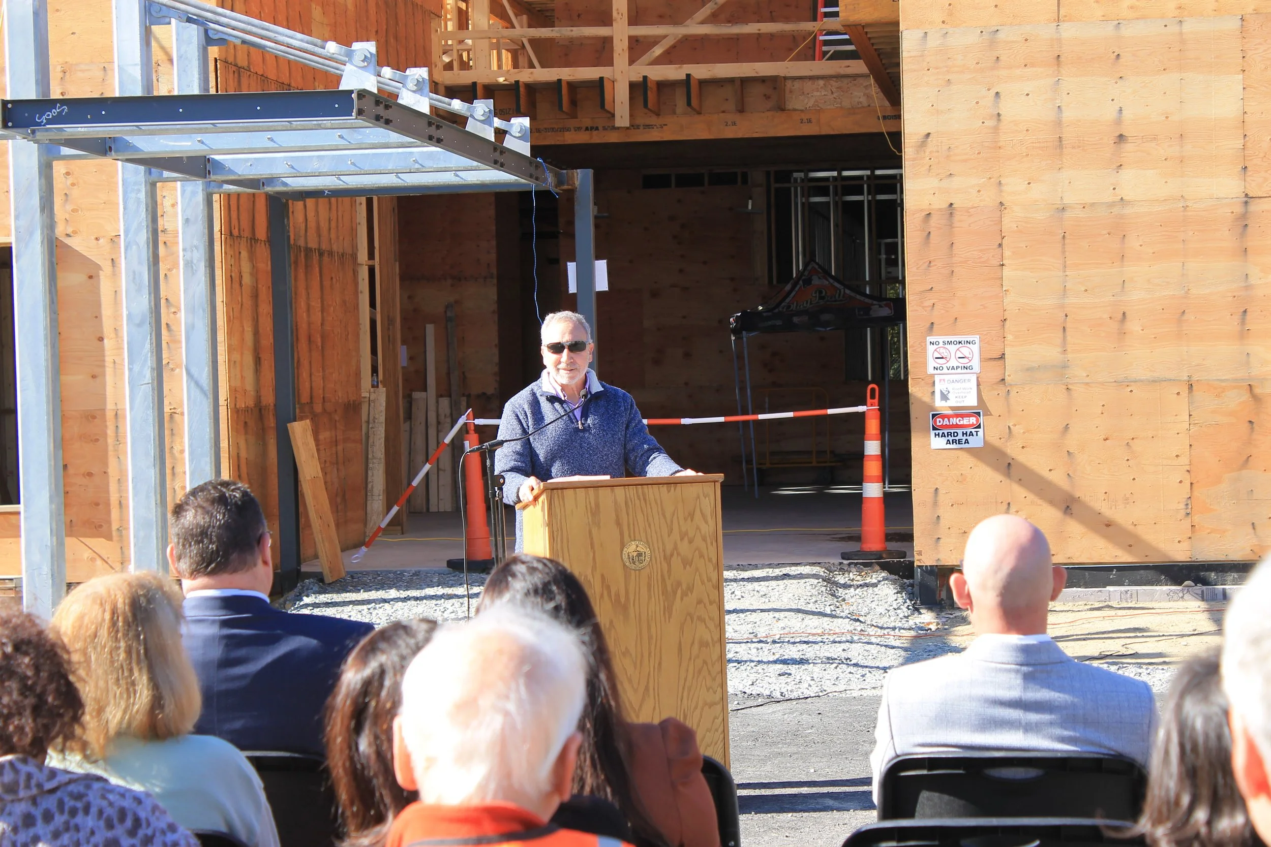 Man with gray hair and sunglasses speaks at a wooden podium before the wood-framed ReCAL building, with seated attendees in the foreground.