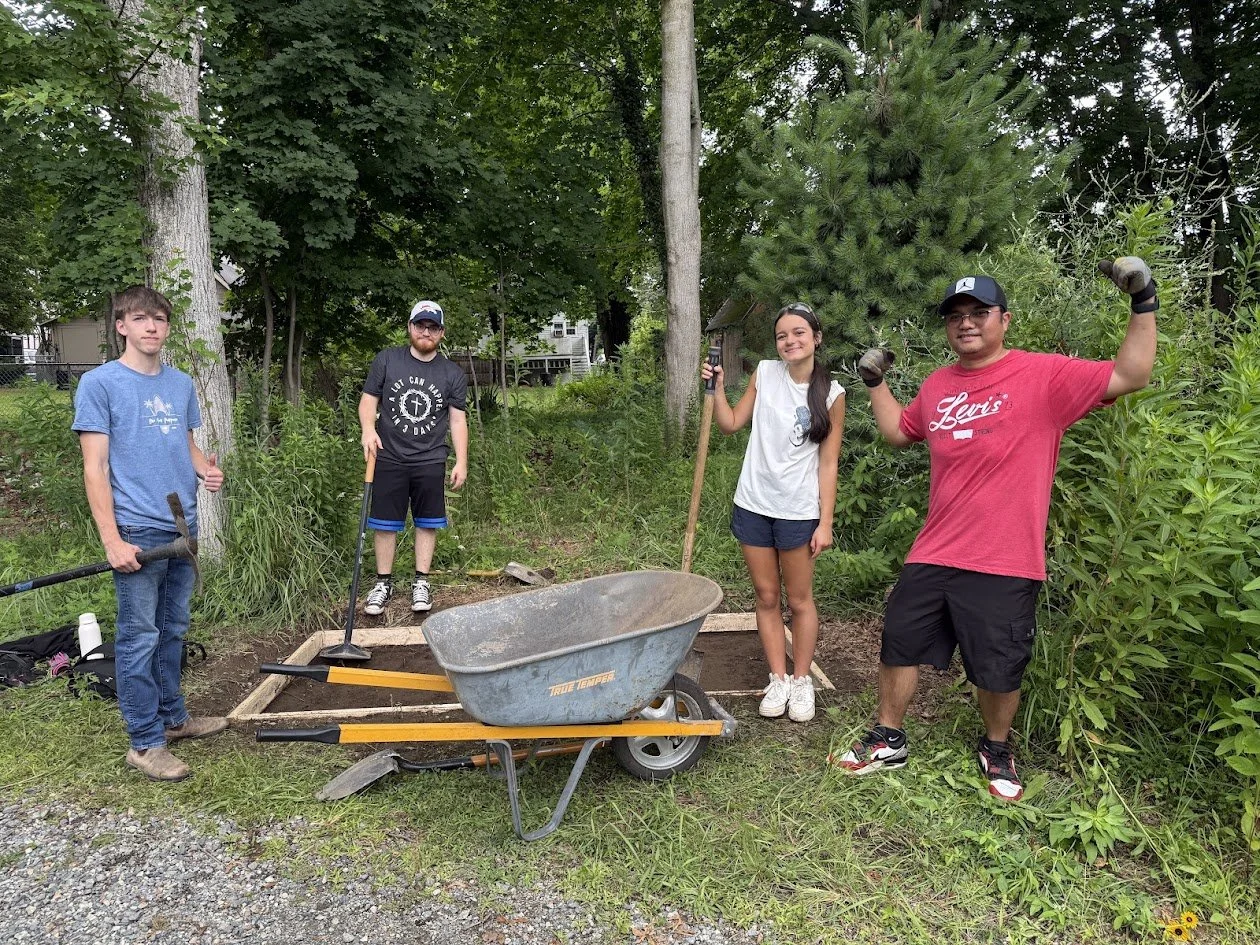 Four volunteers stand behind a wheelbarrow and a wooden form on the ground, holding shovels and rakes.