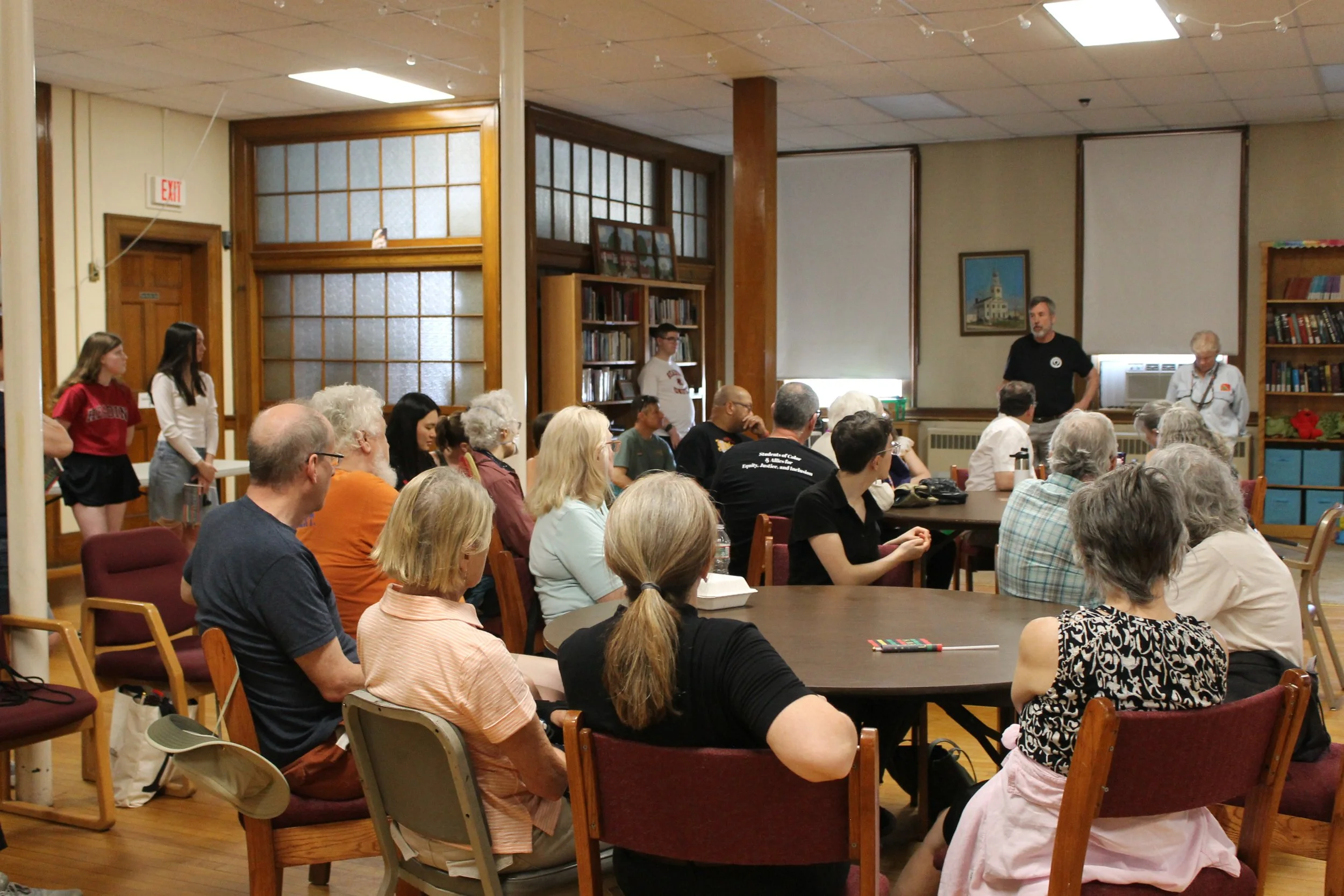 A group of people seated around tables in a wood-paneled community room, listening to a man speaking at the front near bookshelves.