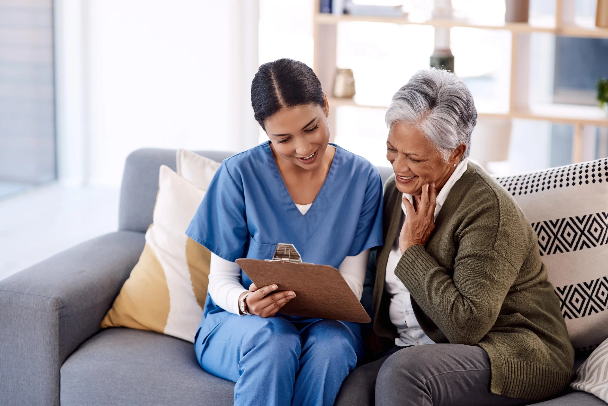 A healthcare worker in blue scrubs shows a clipboard to an elderly woman with gray hair, sitting on a gray sofa, both smiling and sharing a joyful moment.