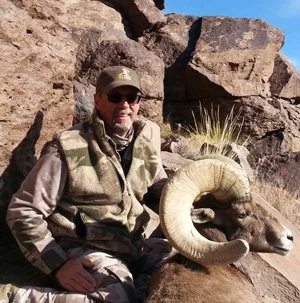 Man in outdoor clothing with sunglasses and a cap sitting next to a ram's head, with rocks and desert plants in the background.