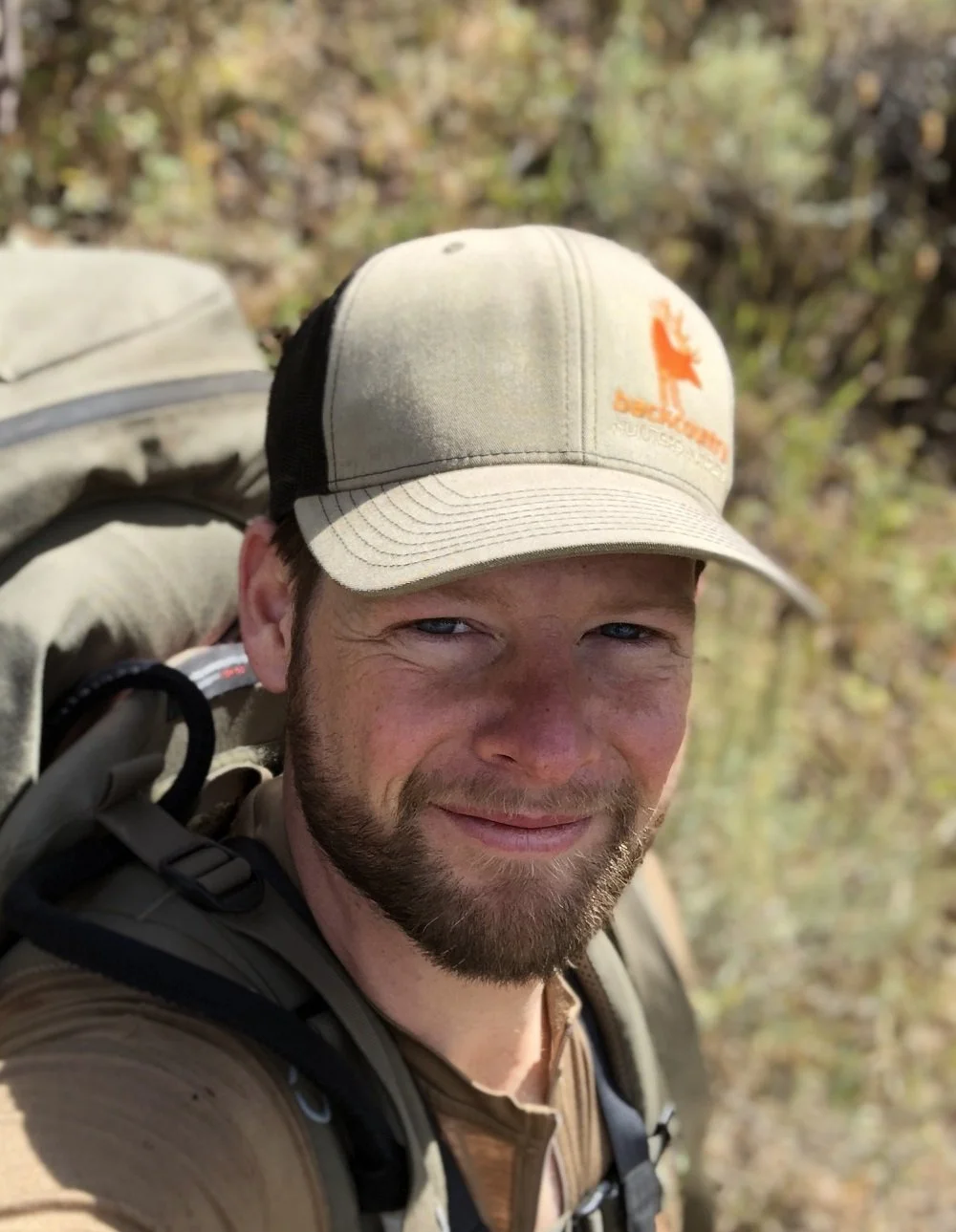A man with a beard taking a selfie outdoors in a wooded area, wearing a beige cap and backpack.