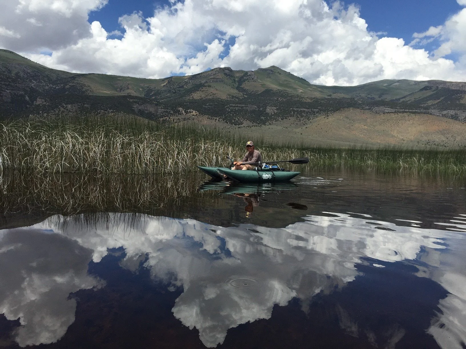 A person sitting in a kayak on a calm lake with mountains and a partly cloudy sky in the background.