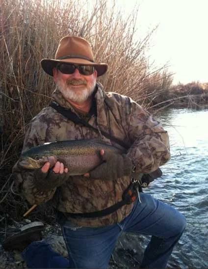 A man wearing a hat, sunglasses, and camouflage clothing is holding a large fish near the water's edge with reeds in the background, smiling.
