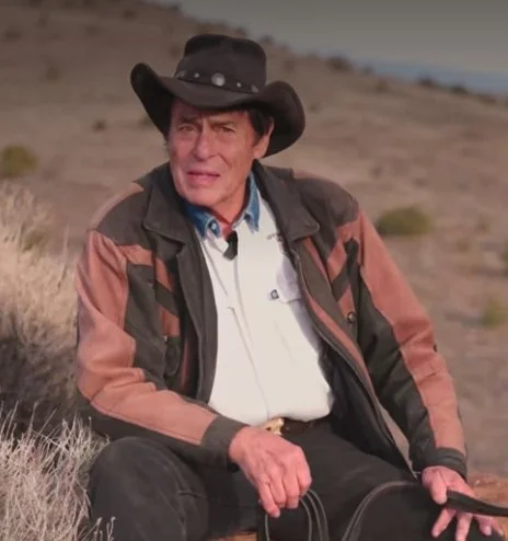 Older man in cowboy hat and outdoor jacket sitting in a high desert sage brush prairie.