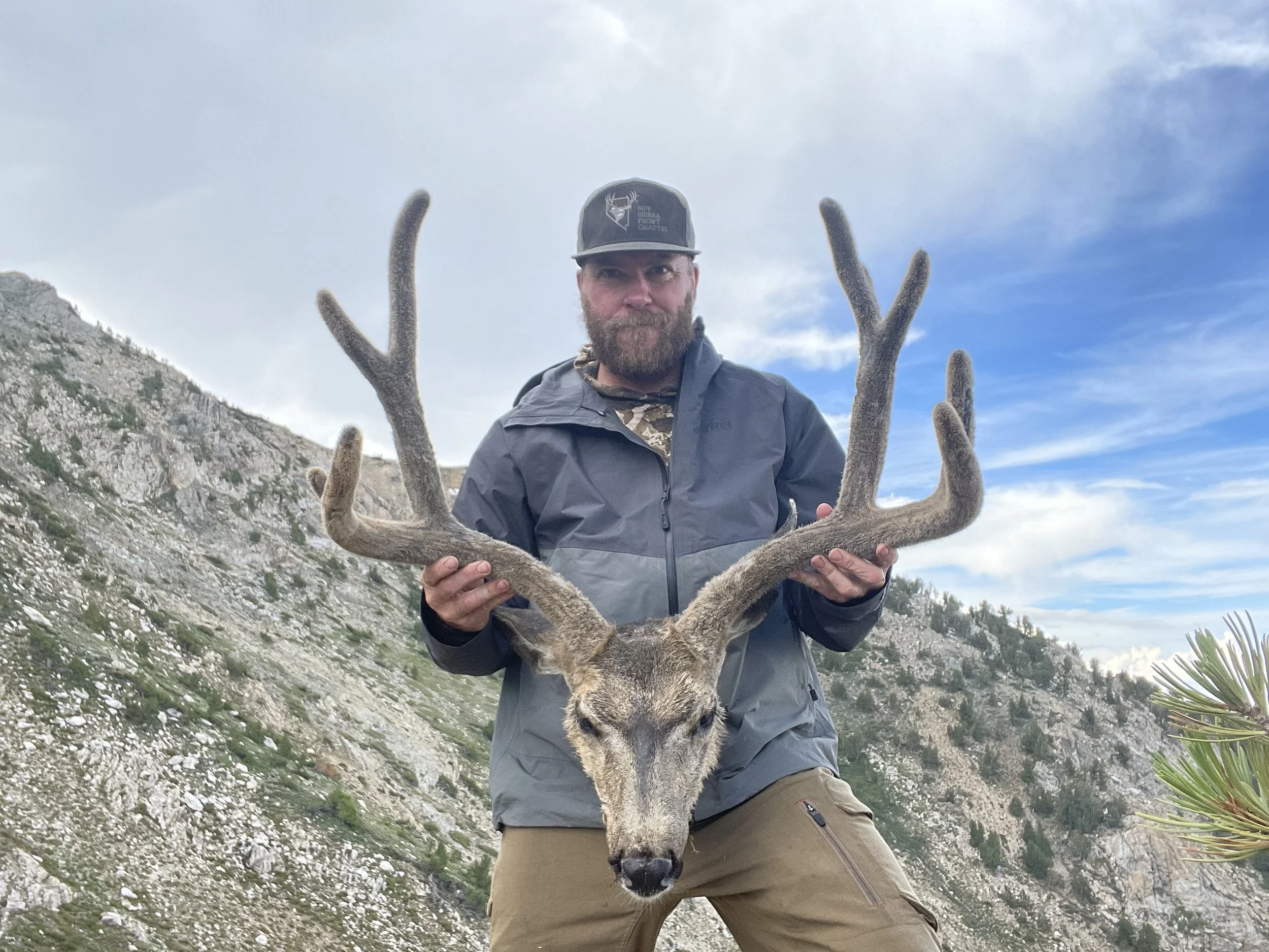 A man with a beard wearing a cap and outdoor jacket holding a large deer antlered skull in a mountain landscape with rocky terrain and sparse vegetation.