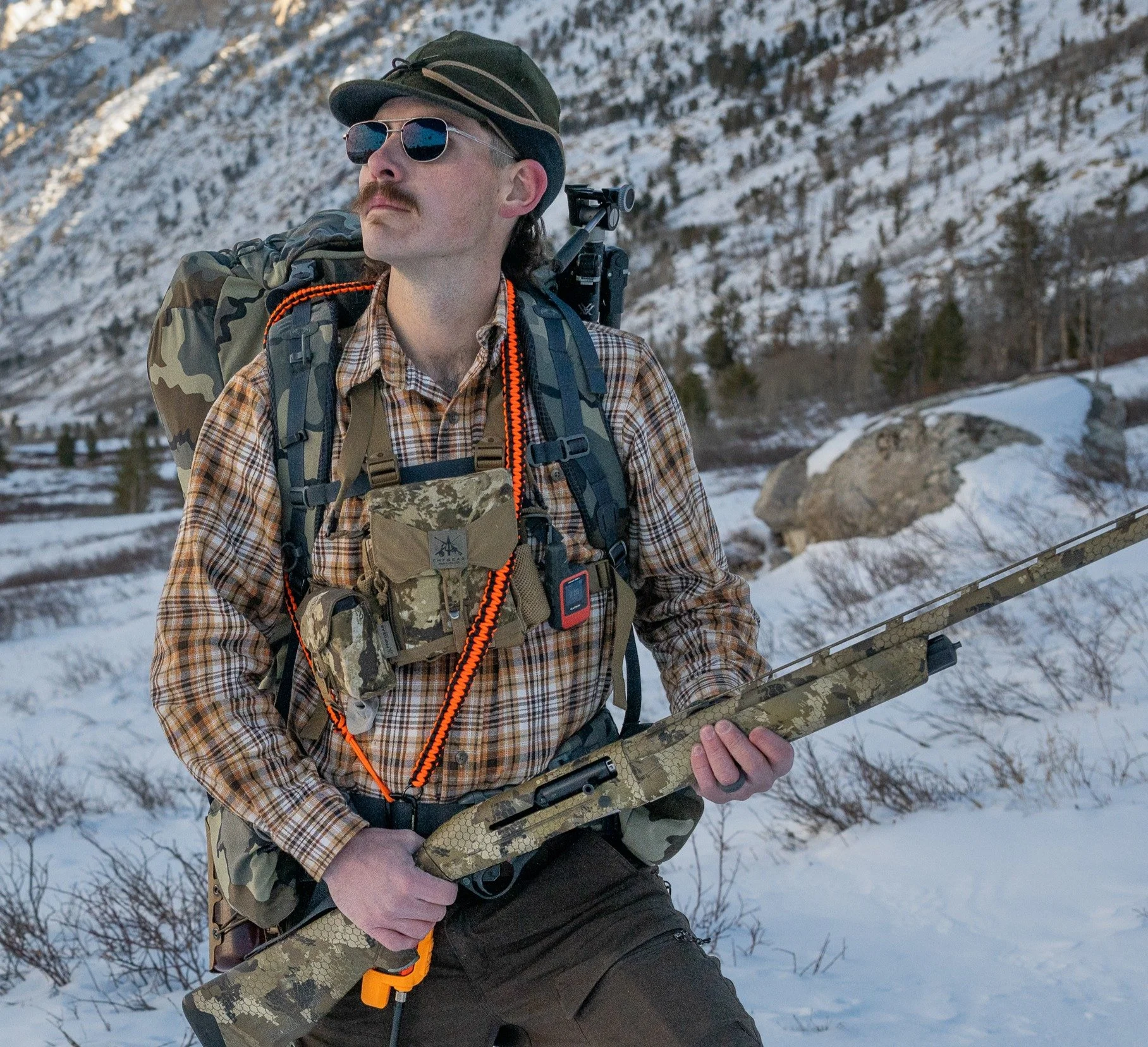Man in camouflage gear with backpack and sunglasses holding a rifle in snow-covered mountainous area.
