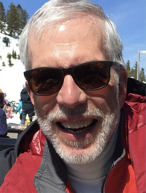 Close-up of a smiling older man with a white beard and sunglasses, outdoors in a snowy area with people and trees in the background.