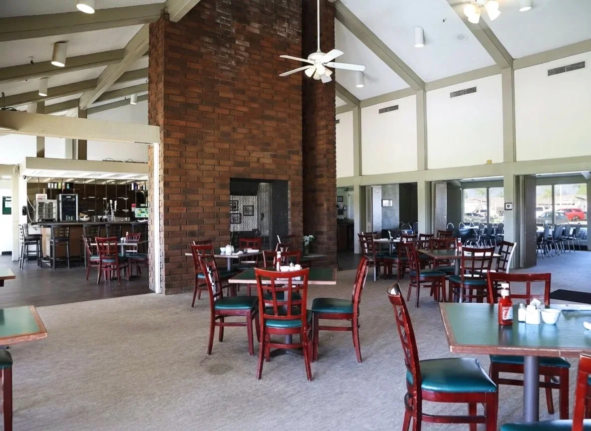 Empty restaurant dining area with redwood tables and chairs, a brick fireplace, and large windows letting in natural light.