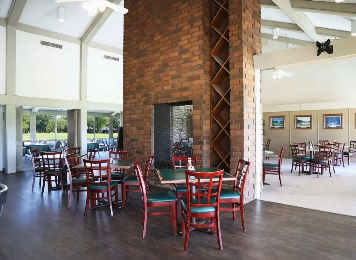 Interior of a restaurant with wooden floors, brick column and tables with chairs, large windows showing outdoor view, and framed pictures on the wall.