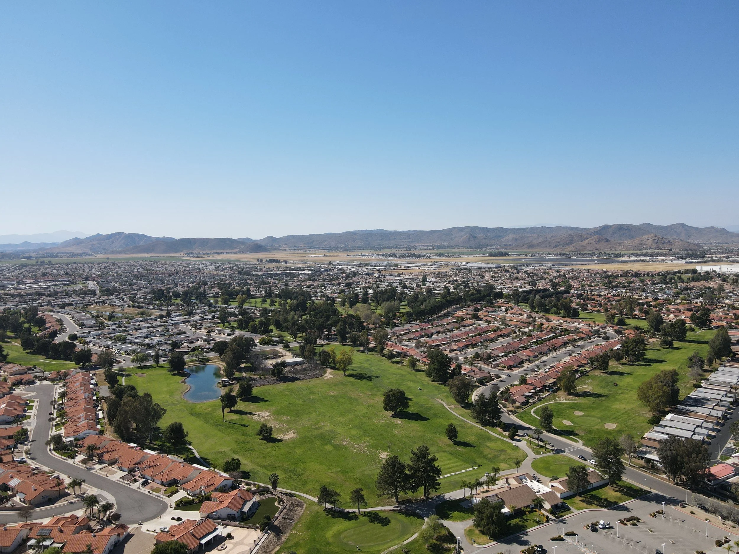 Aerial view of a suburban neighborhood with houses, a park with a pond, Seven Hills Golf Club's course, and mountains in the background under a clear blue sky.