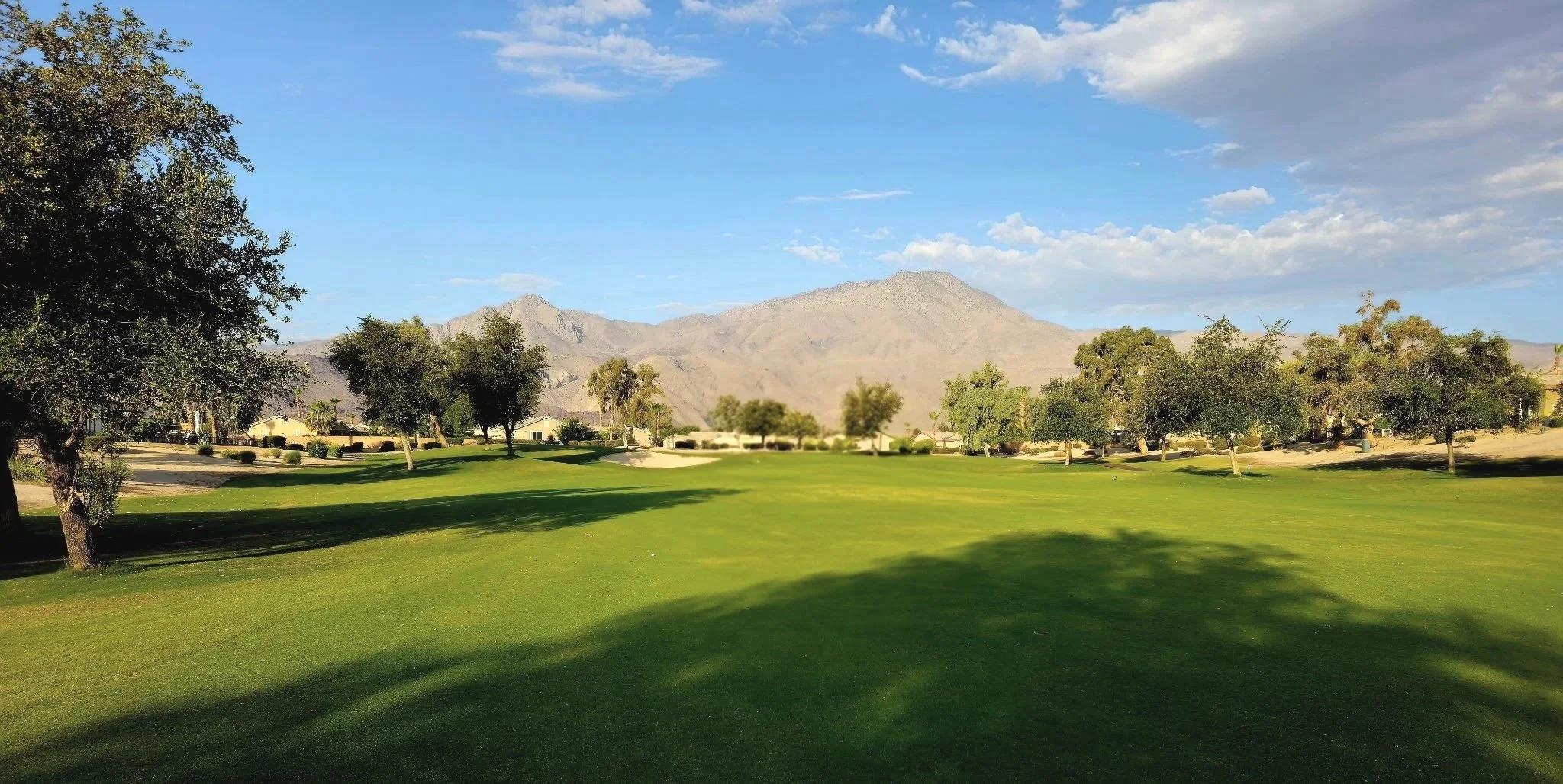 A scenic view of Seven Hills Golf Club's course with lush green grass, scattered trees, and mountains in the background under a partly cloudy sky.