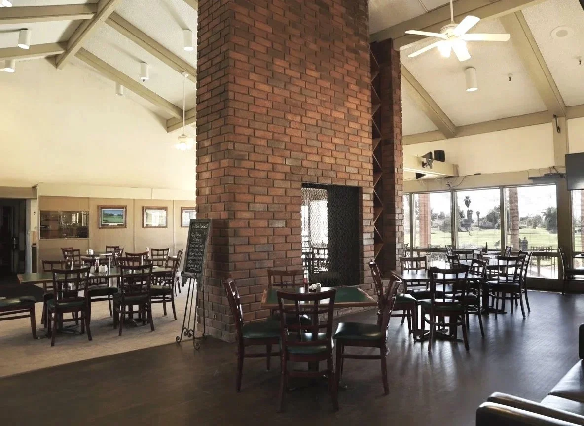Interior of Seven Hills Golf Club restaurant with wooden tables and chairs, large windows with a view of a golf course, a brick fireplace with a chimney, ceiling fans, and wall art.