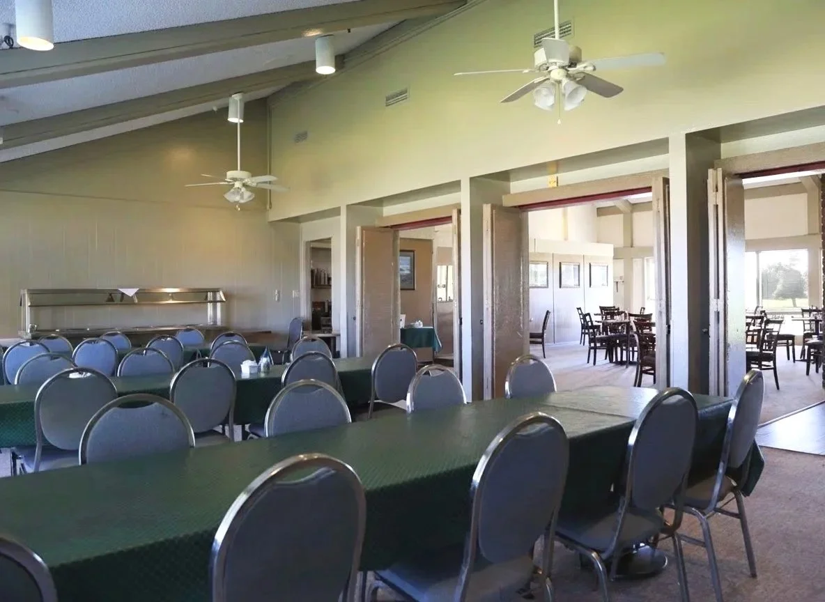 A dining area with multiple tables covered with green tablecloths, surrounded by gray chairs. There are ceiling fans and large windows letting in natural light, and a partial view of another room with additional seating.