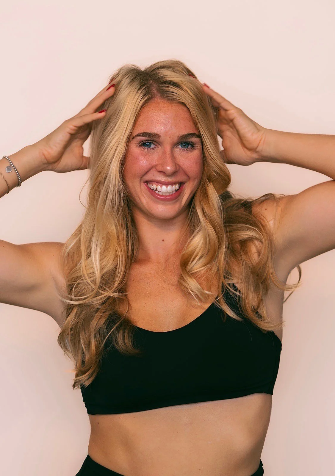 Smiling woman with long blonde hair and blue eyes, wearing a black sports bra, holding her hair with both hands, against a plain white background.