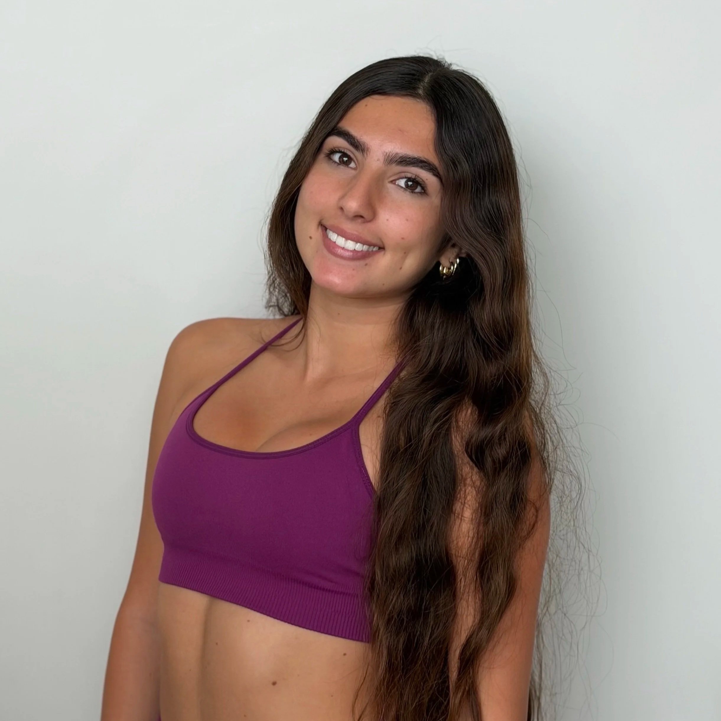 Young woman with long brown hair and gold hoop earrings smiling in front of a plain wall, wearing a purple sports bra.