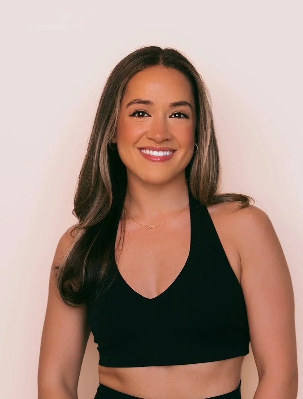A young woman with long wavy brown hair wearing a black sports bra and a delicate gold necklace, smiling against a plain light-colored background.