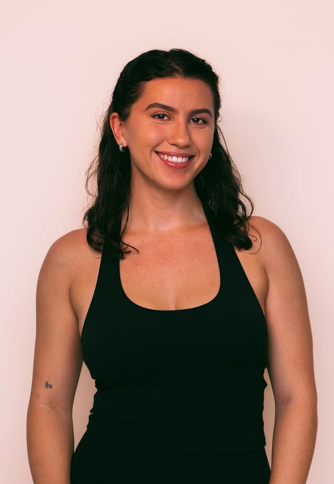 Smiling young woman with dark, wavy hair in a black tank top, standing against a plain light background.