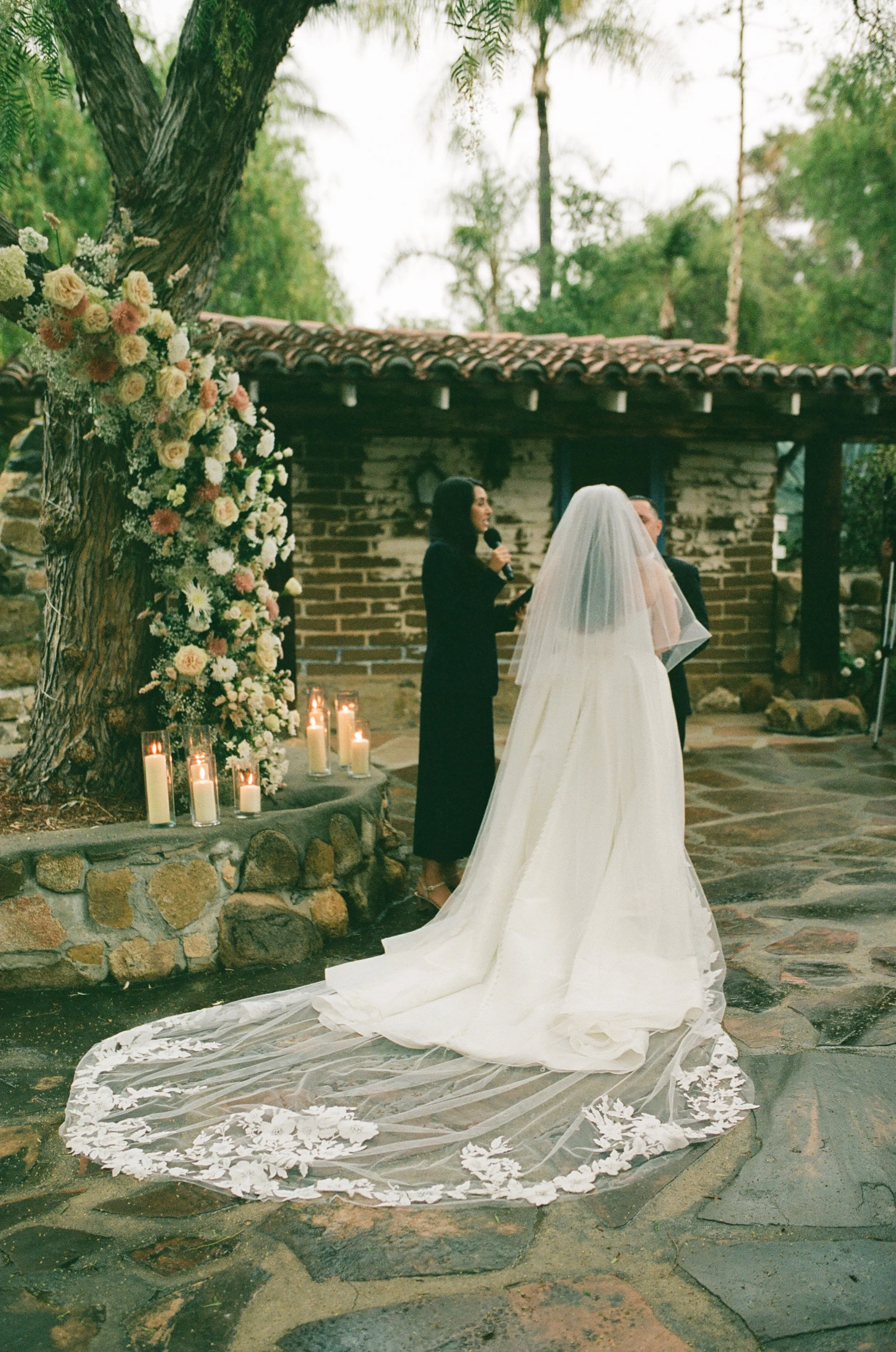 A bride and groom exchanging vows during an outdoor wedding ceremony with a woman officiant, lit candles, and floral decorations, set against a rustic building and trees.