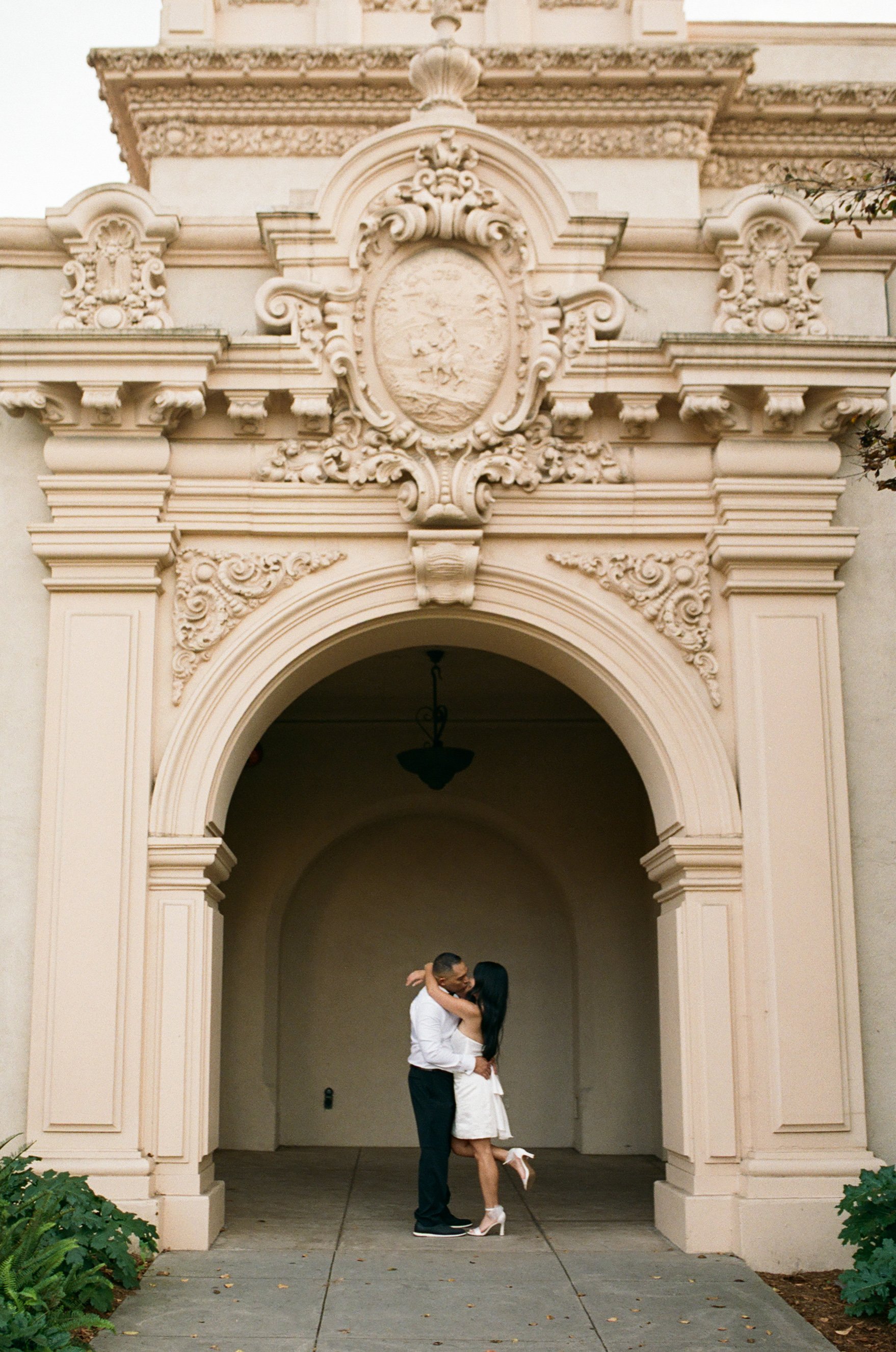 A couple kissing under an ornate archway of a historic building with decorative stonework and a crest at the top.
