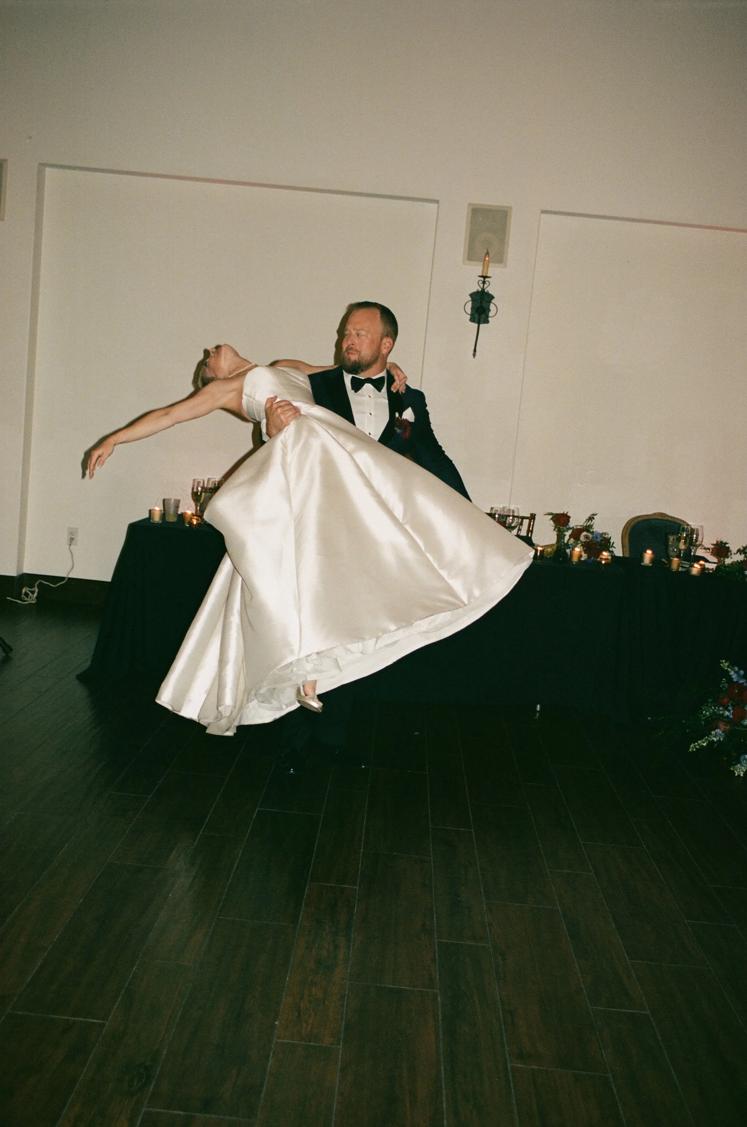 A groom in a tuxedo carrying a bride in a white wedding gown during their wedding reception, with a decorated table and candles in the background.