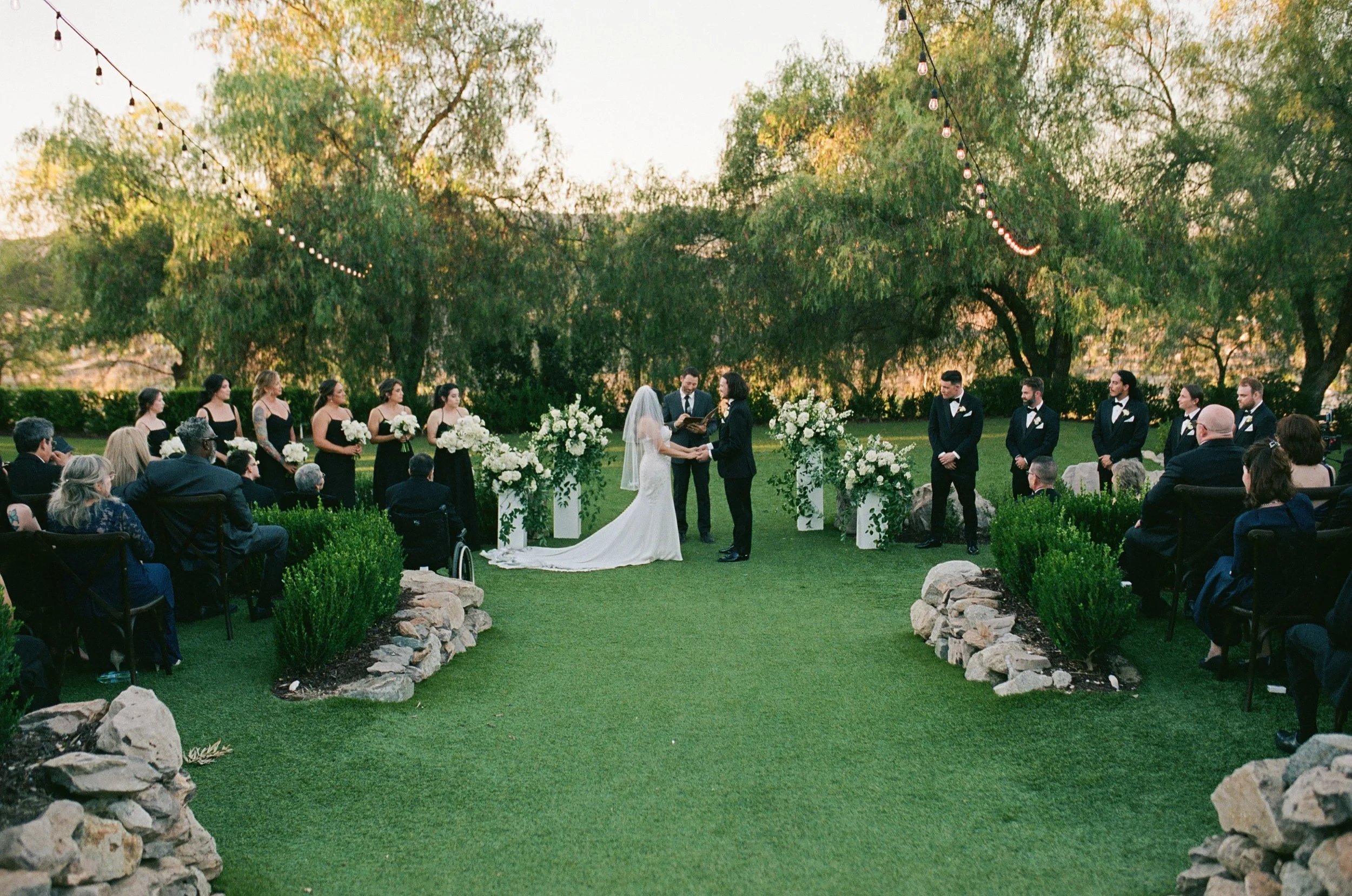 Outdoor wedding ceremony with a bride and groom facing each other, surrounded by bridesmaids in black dresses and groomsmen in tuxedos, under string lights and large trees, with guests seated on either side.