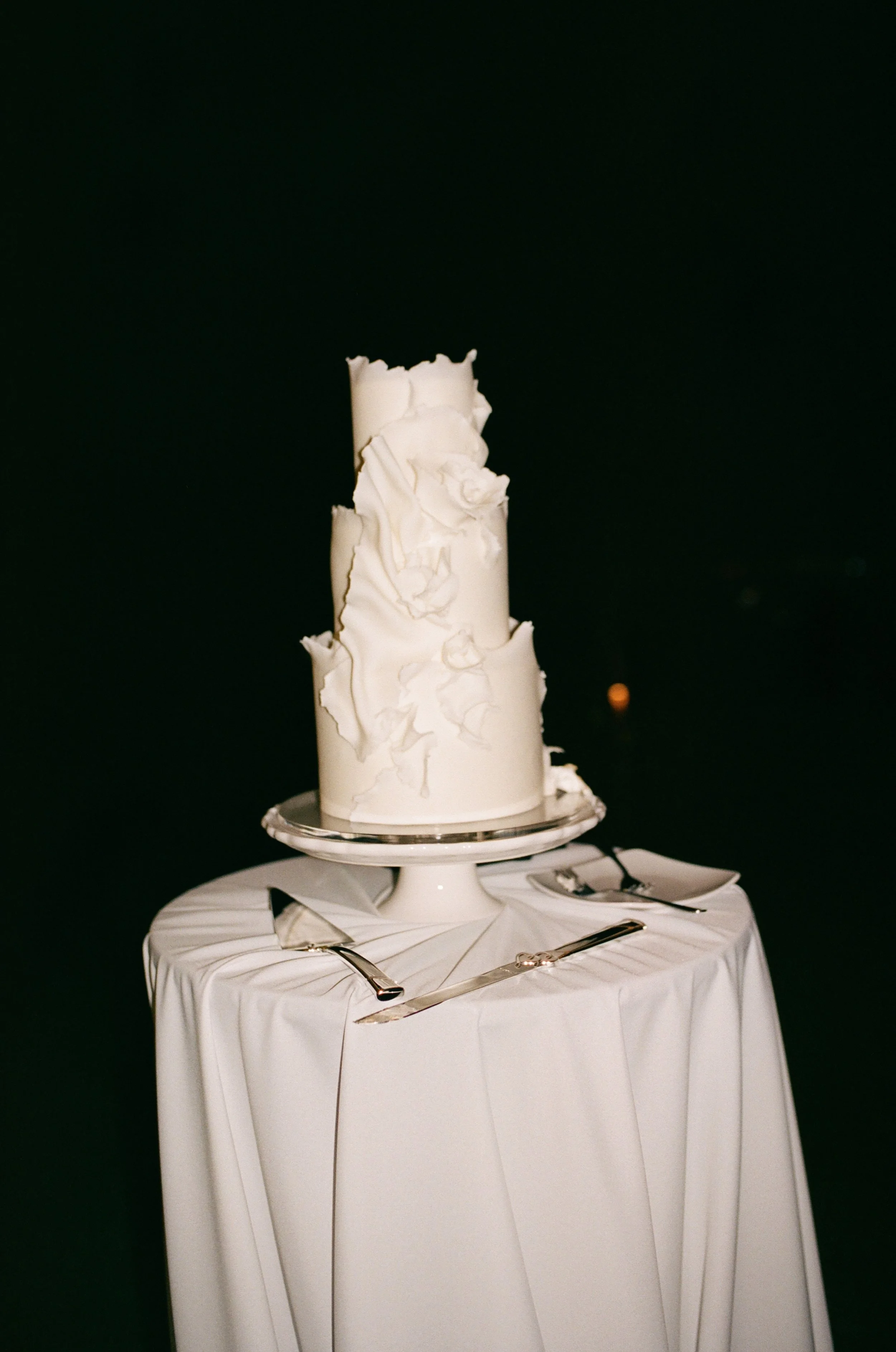 A tall, white, three-tiered wedding cake with textured icing and floral accents, placed on a cake stand on a round white table with a white tablecloth. Cake knives and servers are arranged around the cake on the table.