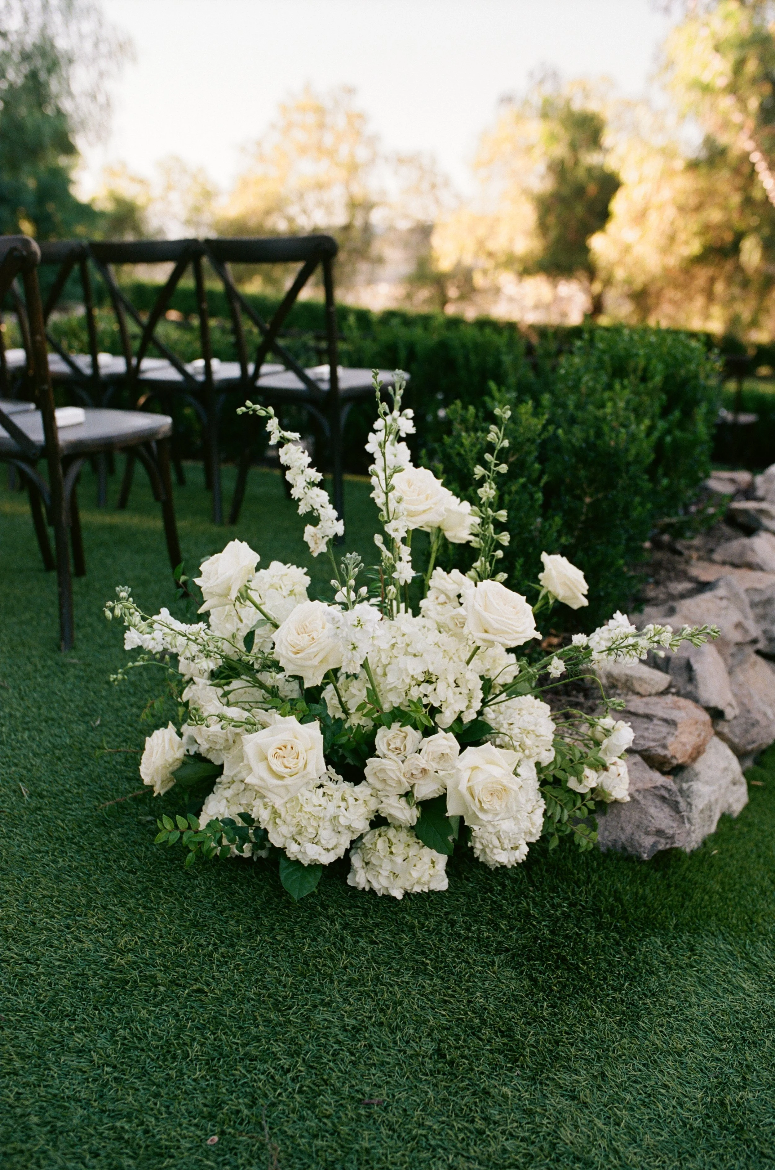 A bouquet of white roses and hydrangeas on green grass, with black chairs and trees in the background.