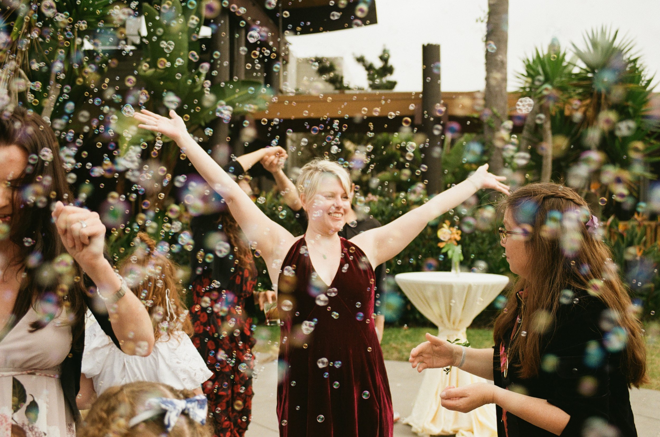 A group of women celebrating outdoors with bubbles, dancing, and smiling, in a garden setting with trees, plants, and flowers.