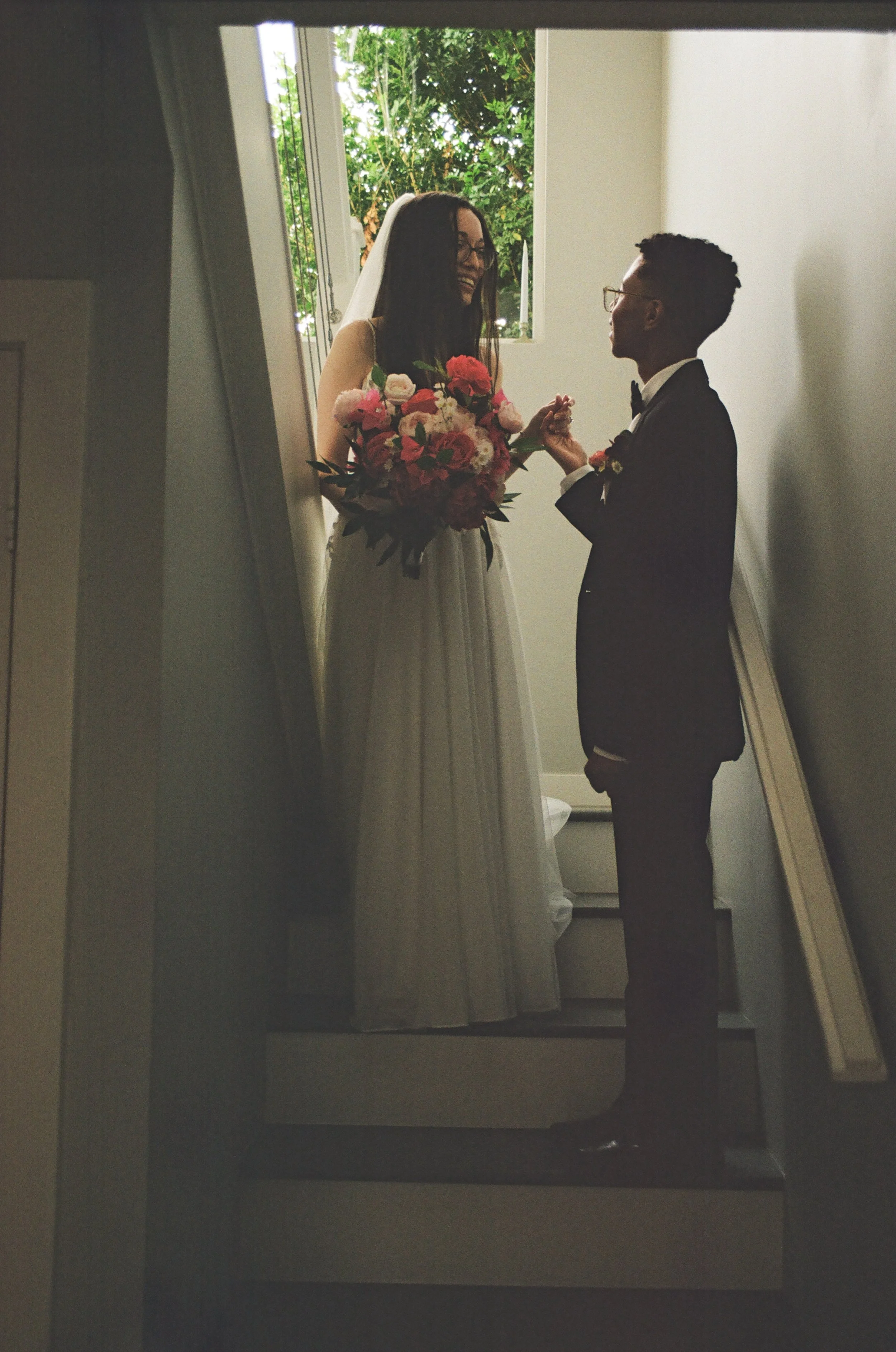 A bride in a white dress holding a bouquet of pink and red flowers standing on a bathroom staircase, smiling at a groom in a black tuxedo with glasses, holding her hand, inside a house with a window showing greenery outside.