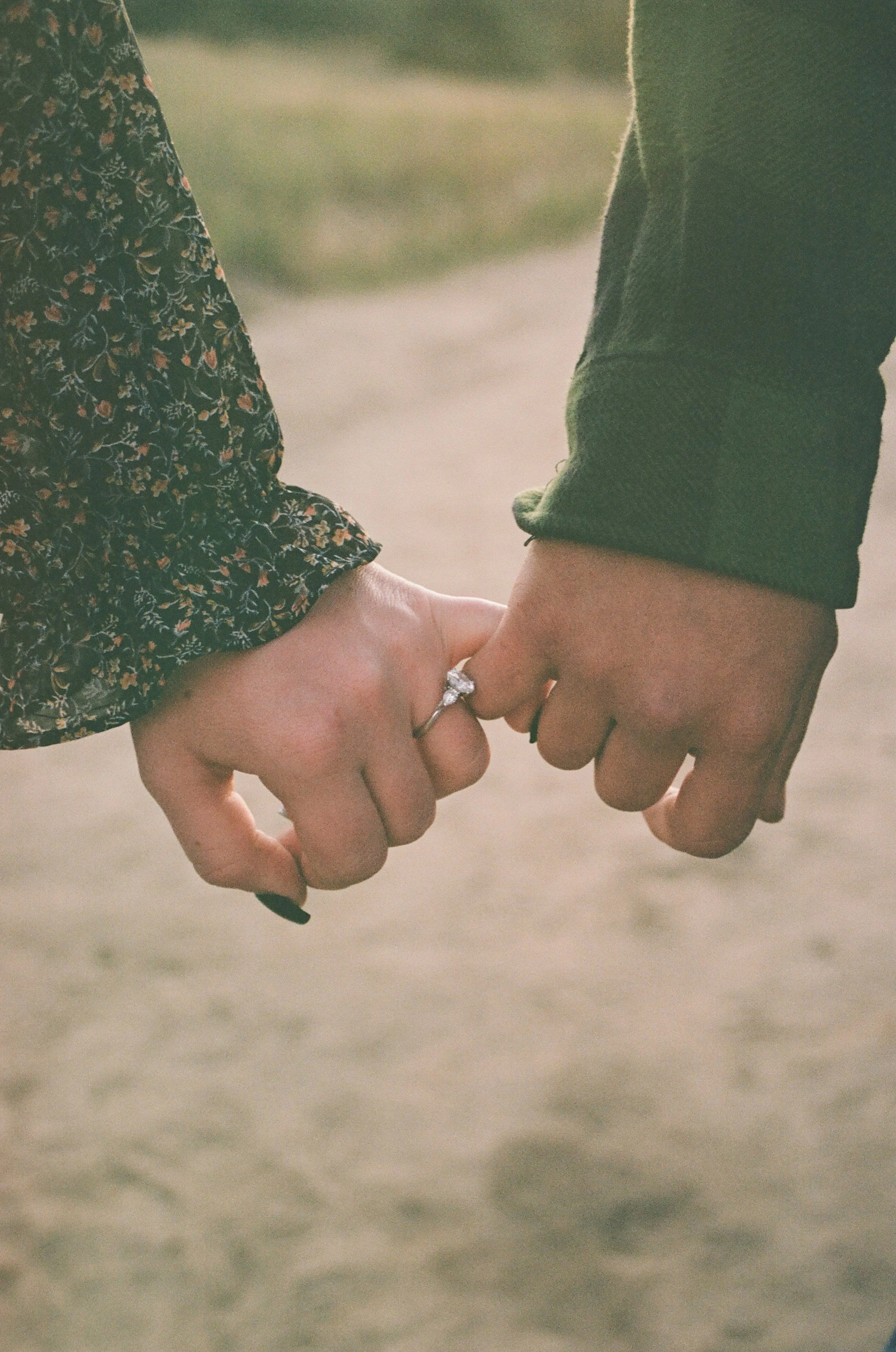Close-up of a couple holding hands, with wedding rings visible, outdoors during daytime.