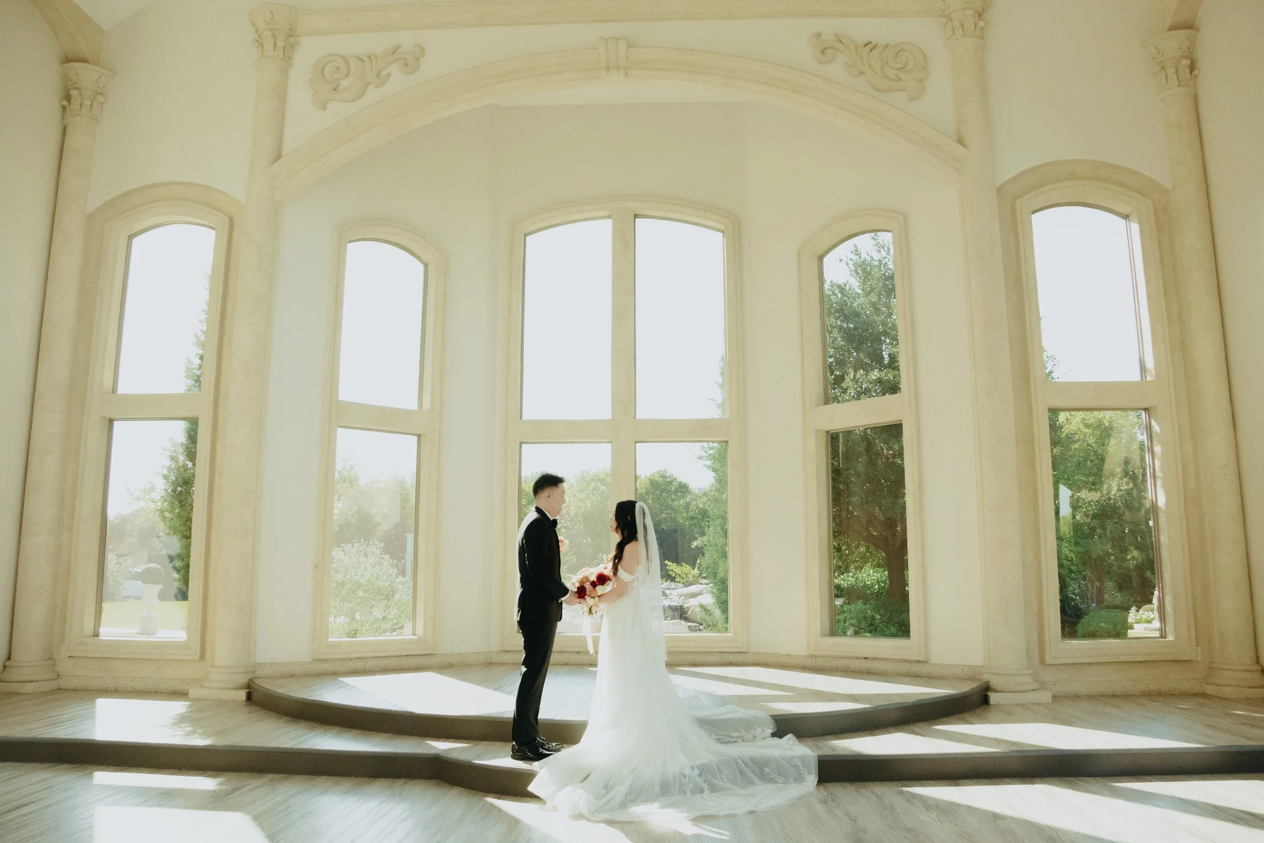 A bride and groom standing inside a bright, elegant room with large arched windows. The couple faces each other, holding hands, with the bride holding a bouquet of flowers. The room features ornate architectural details and natural light streaming through the windows, with greenery visible outside.