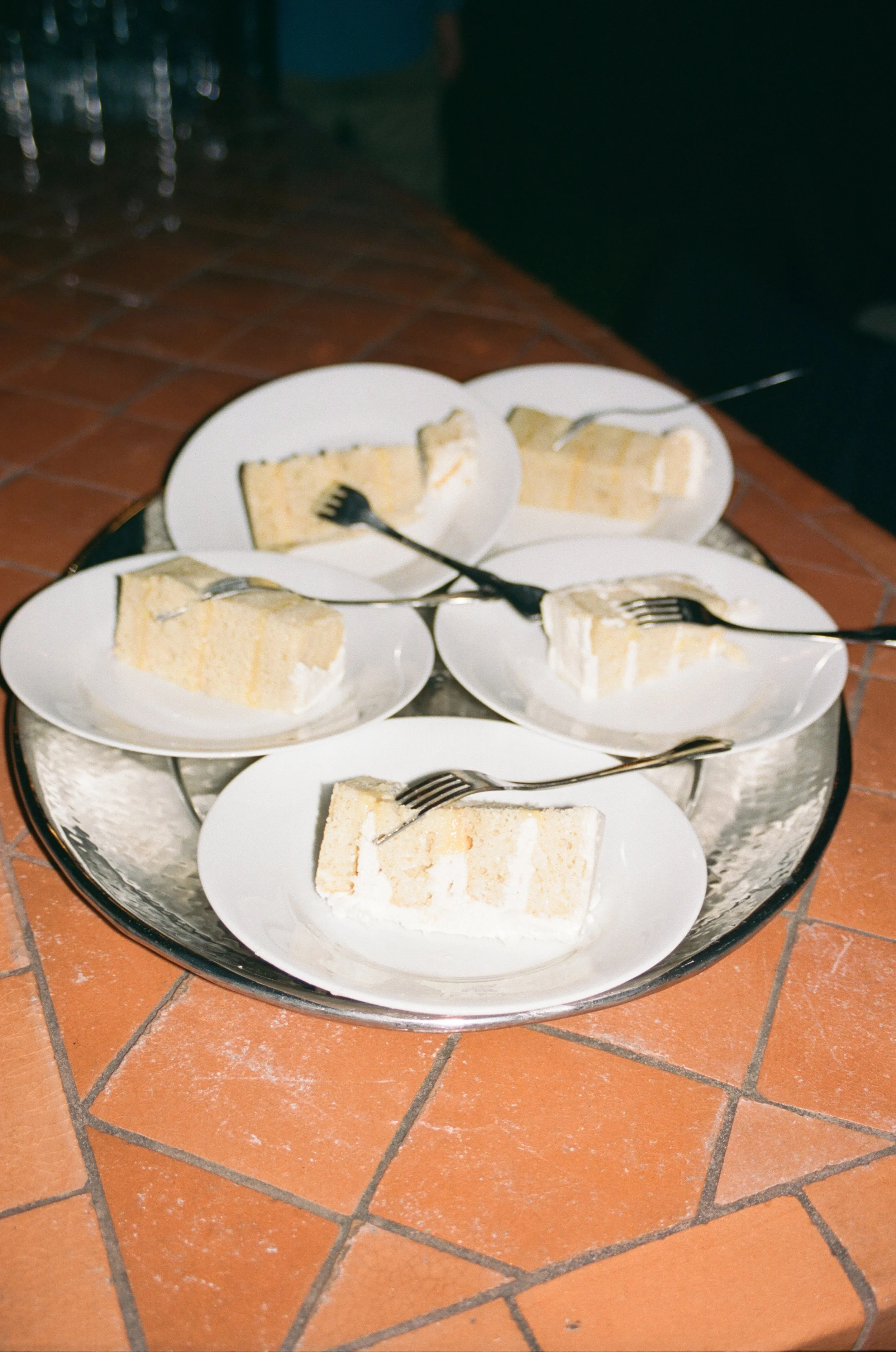 Four slices of white cake with white frosting on separate small white plates, each with a fork, arranged on a large silver tray on a brick floor.