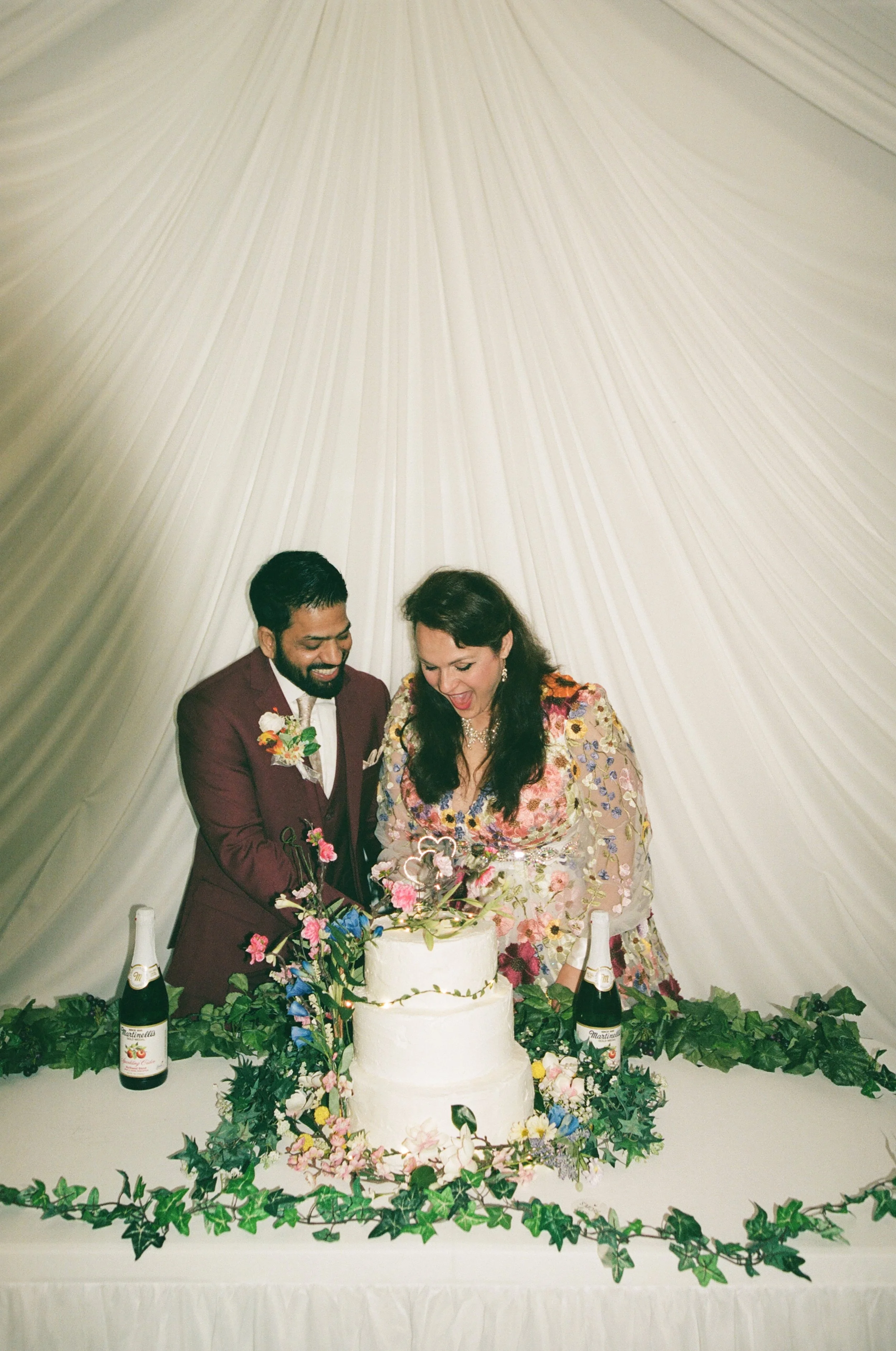 A newlywed couple cutting their wedding cake, surrounded by greenery and flowers, with champagne bottles on the table.