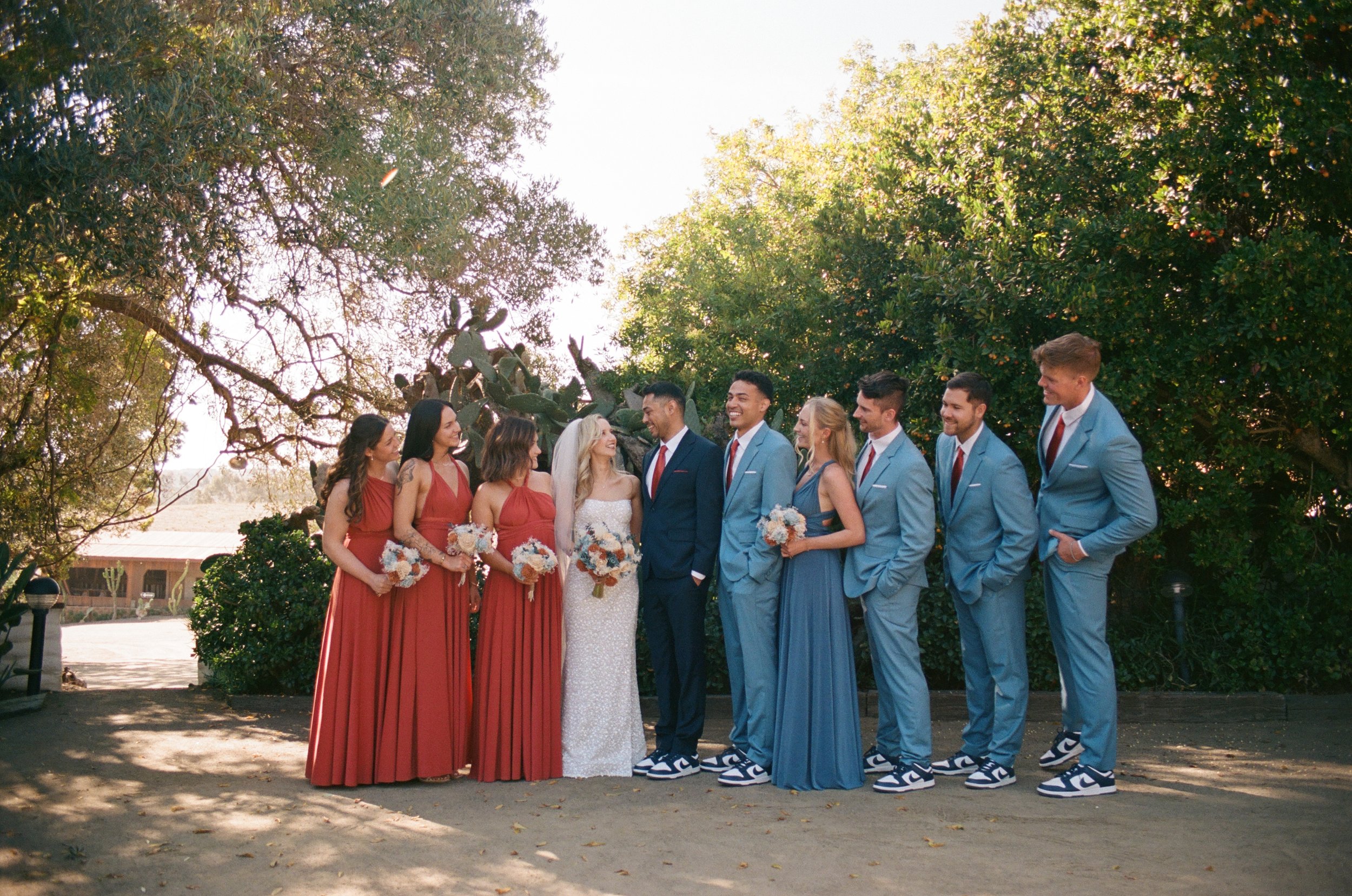 A wedding party with bridesmaids wearing red dresses and holding bouquets, groomsmen in light blue suits and sneakers, standing outdoors under trees during daytime, smiling and looking at each other.
