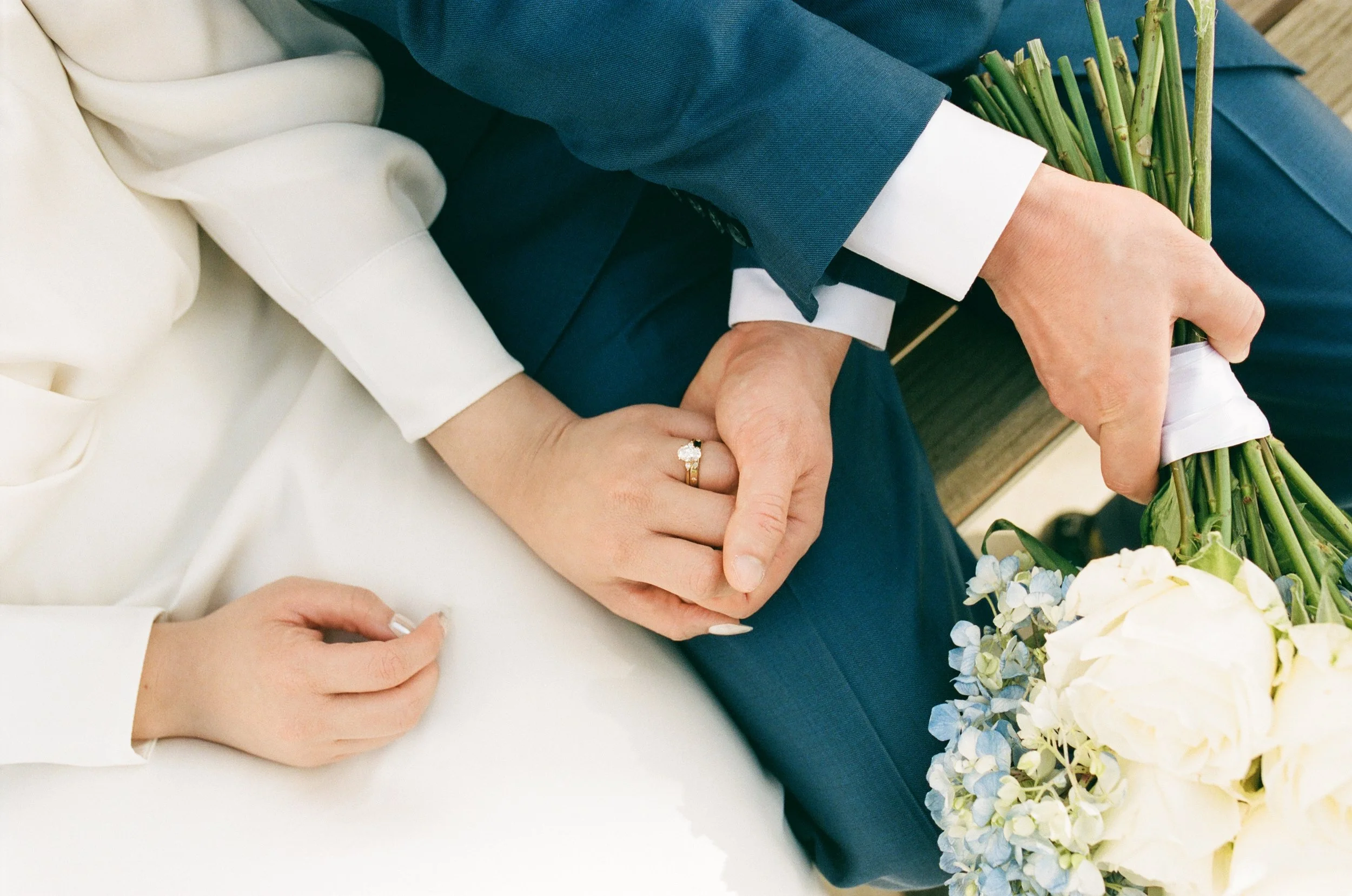 Close-up of a couple holding hands during a wedding ceremony, with the bride wearing a white dress and the groom in a navy suit holding a bouquet of white and pale blue flowers.