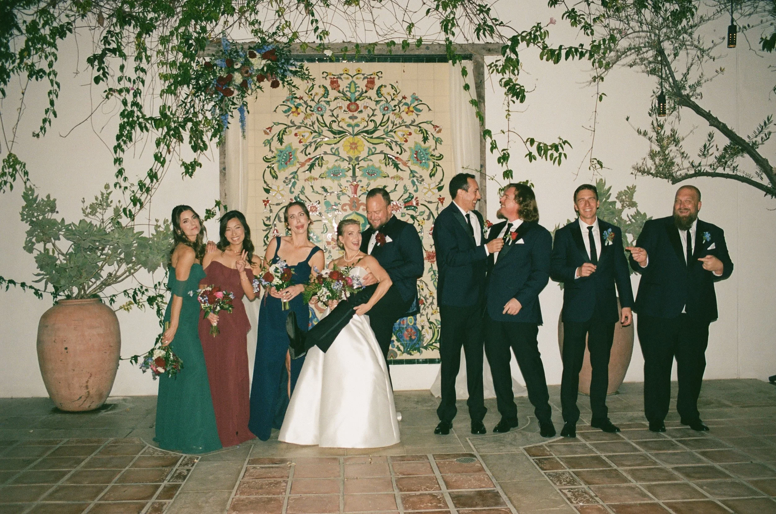 A group of wedding guests dressed in formal attire, standing and celebrating indoors, with a decorative tapestry and plants in the background.