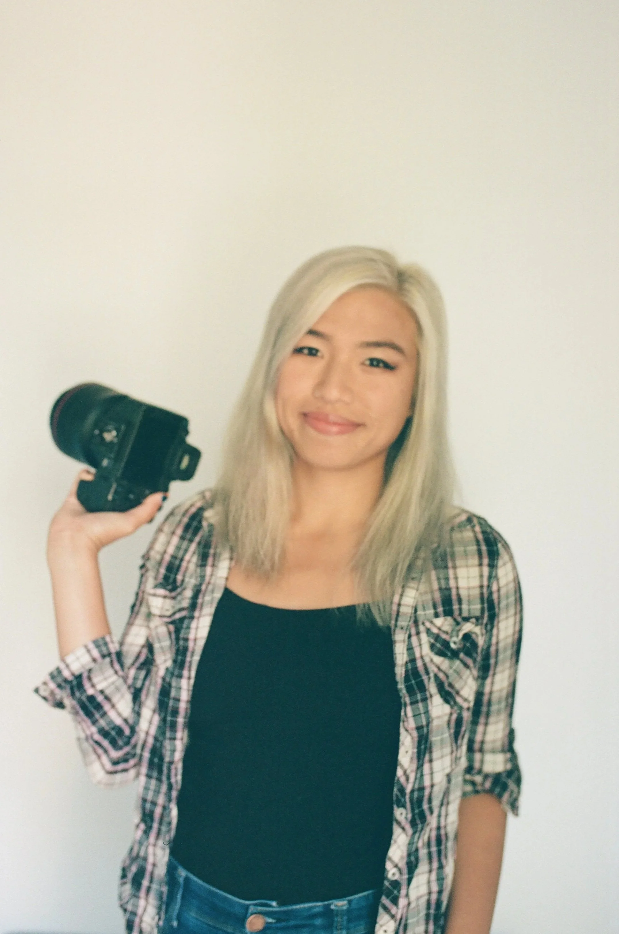 A young woman with blonde hair, wearing a black top and plaid shirt, smiling and holding a camera in her right hand against a plain white wall.