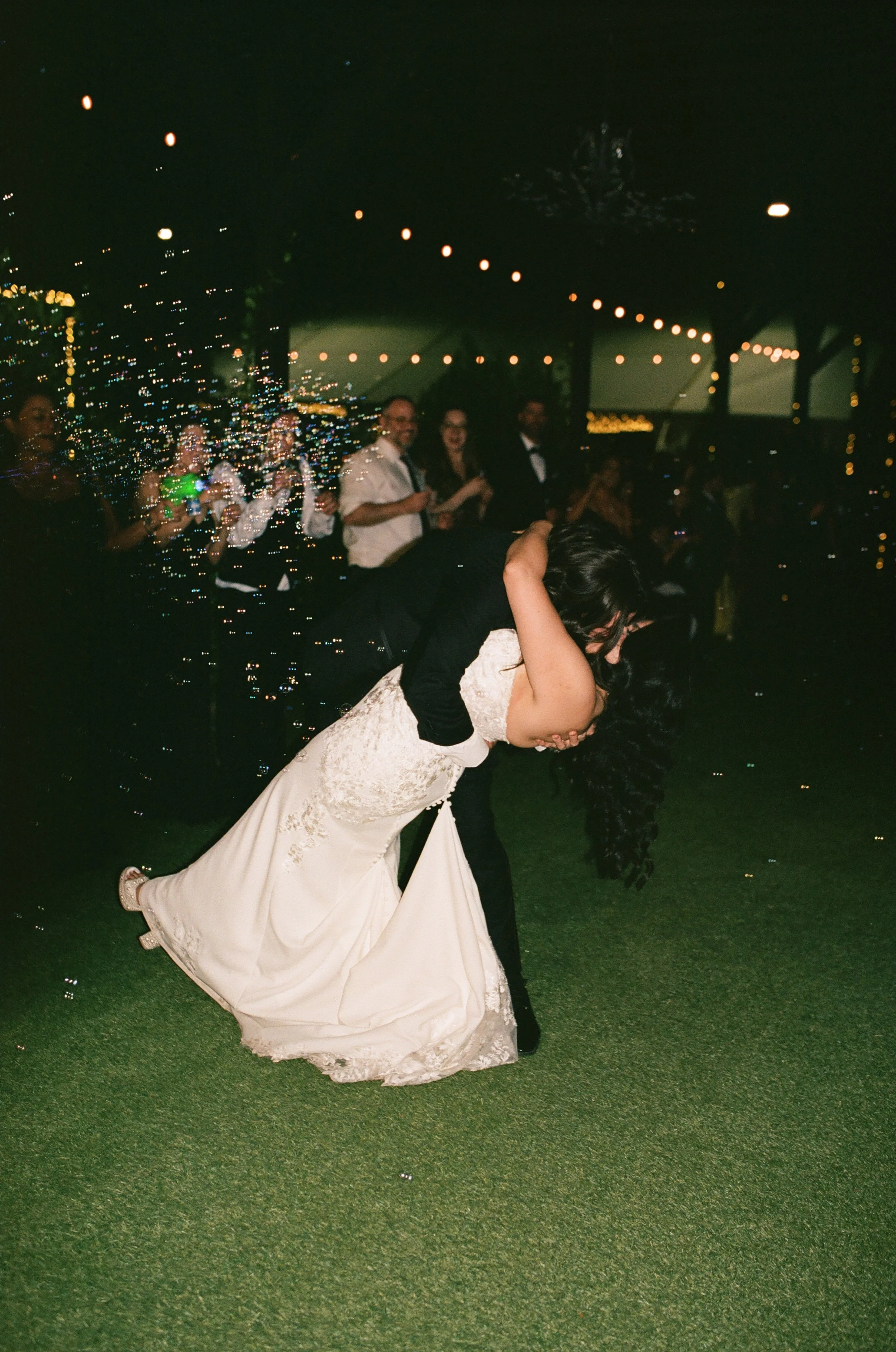 Couple dancing during their wedding reception with guests and string lights in the background.