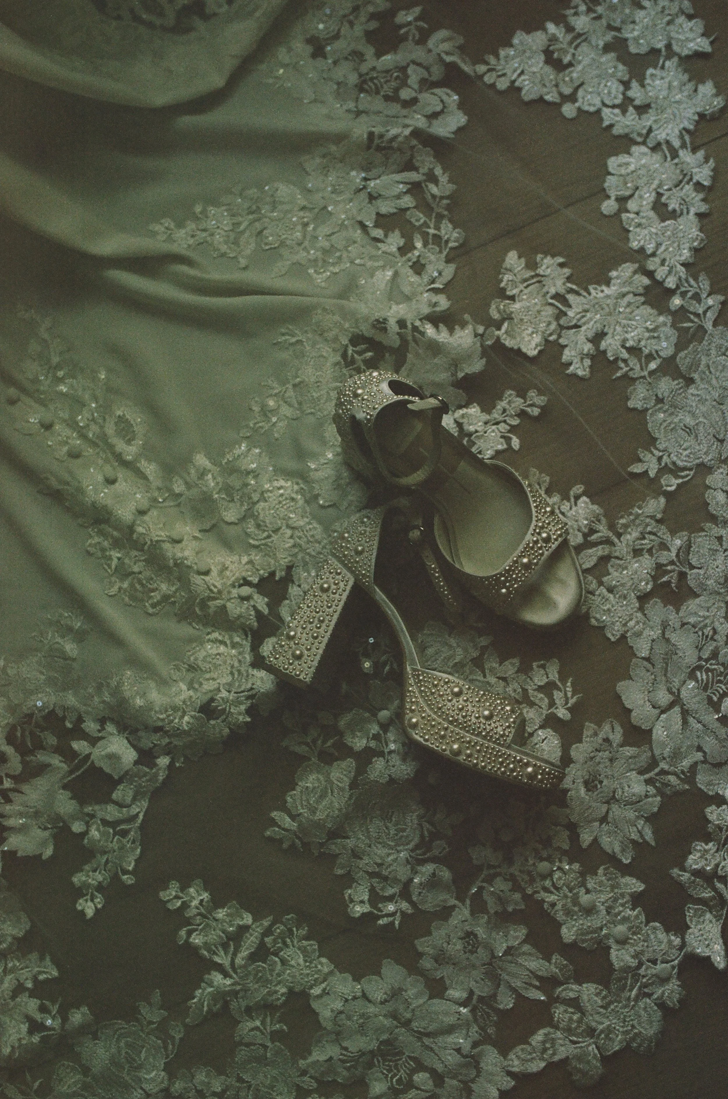 A pair of high-heeled shoes decorated with pearls and rhinestones, resting on a floral lace fabric.