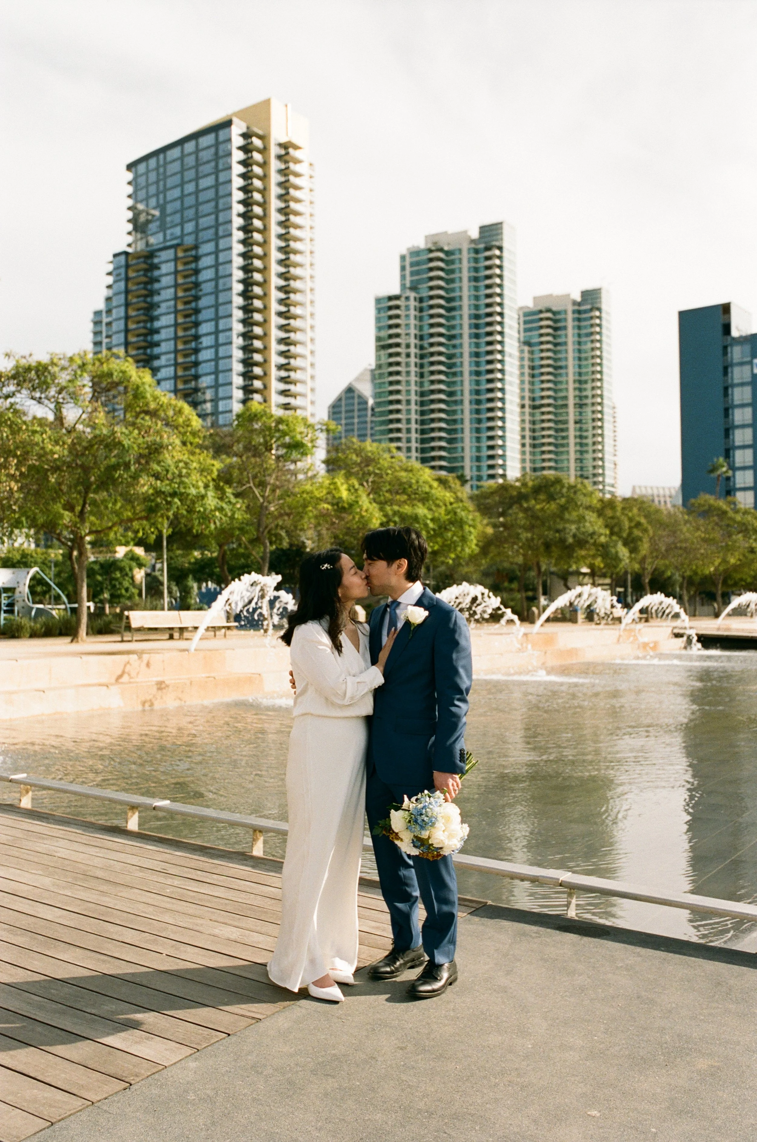 A couple in wedding attire sharing a kiss by a fountain in an urban park, with city high-rise buildings in the background.