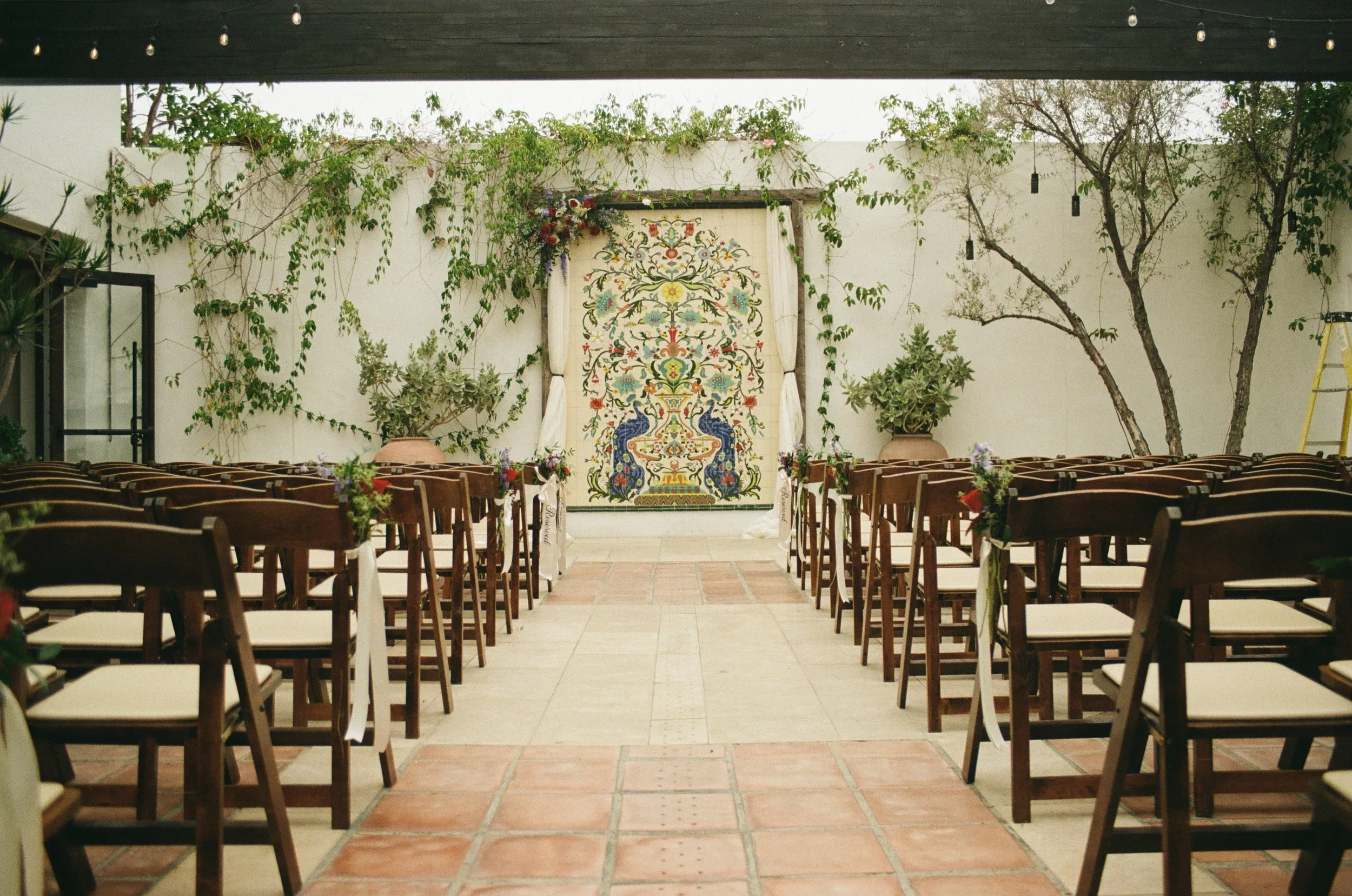 A small outdoor wedding setup with wooden chairs arranged in two rows facing a decorated backdrop on a white wall with greenery. The backdrop features a colorful, intricate tapestry and is framed by white curtains, with plants and trees on either side.