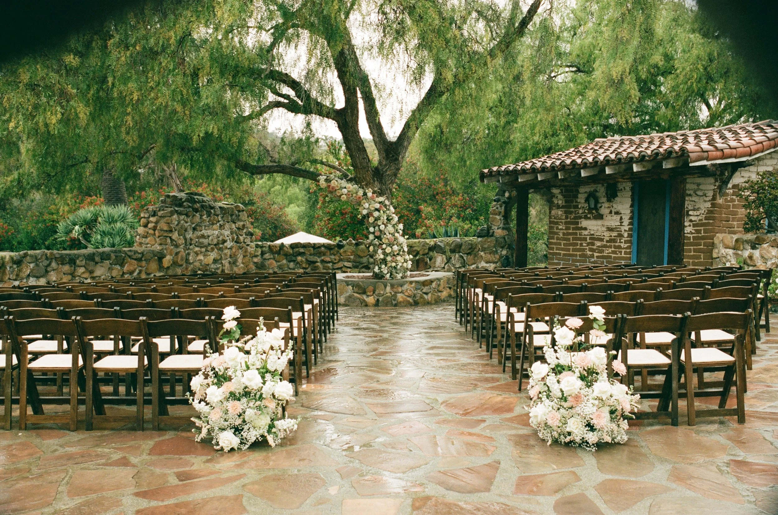 Outdoor wedding ceremony setup with rows of chairs on either side of an aisle adorned with white flowers, beneath a large leafy tree with pink and white flowers, on a stone-paved courtyard surrounded by greenery and a rustic brick building.